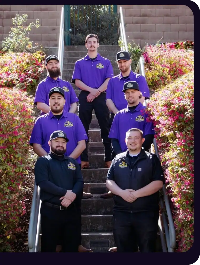 Eight men stand on outdoor steps, seven in purple shirts and one in black, surrounded by bushes with red and yellow foliage.