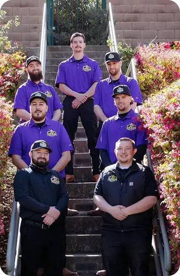 Six men pose on outdoor stairs, five wearing matching purple shirts and hats with logos, and one in a black shirt, all smiling at the camera. Shrubs line both sides of the staircase.