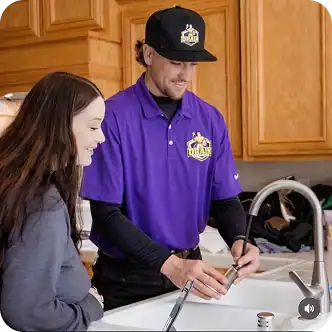 A man in a purple uniform demonstrates fixing a kitchen faucet while a woman watches beside him in a kitchen.